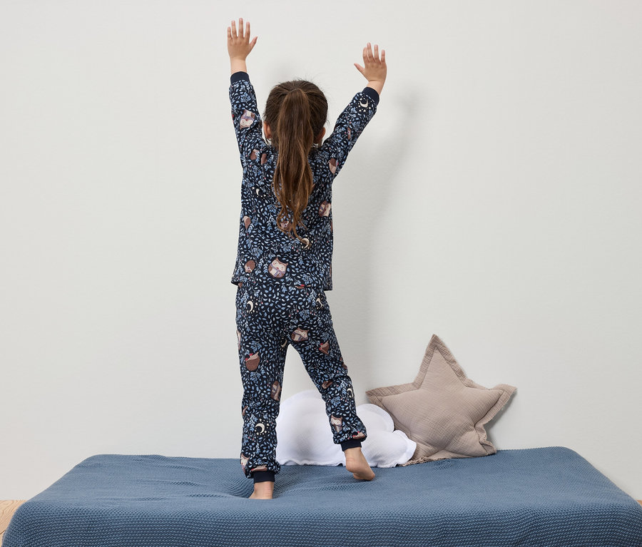Mädchen im dunkelblauen Schlafanzug mit Aufdruck steht mit erhobenen Händen auf einem blauen Bett.