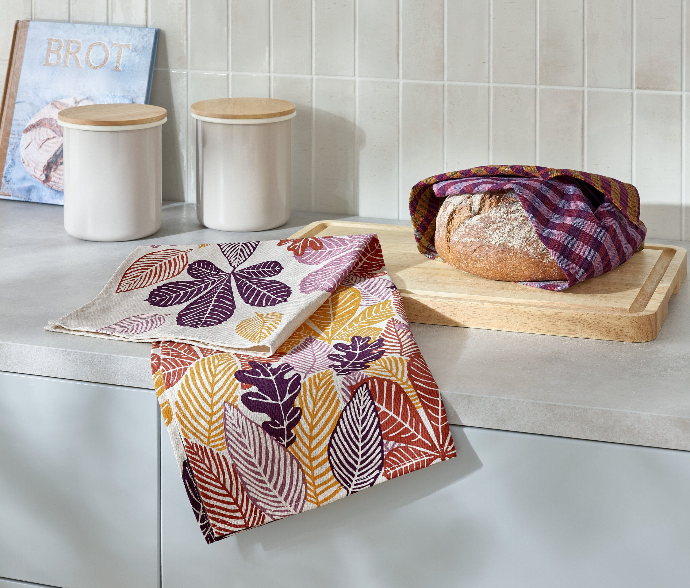 Auf einer Küchentheke liegen ein in ein kariertes Tuch gehüllter Laib Brot auf einem Holzbrett, daneben zwei Geschirrtücher mit Blätterdruck, zwei weiße Dosen und ein Buch mit der Aufschrift Brot.