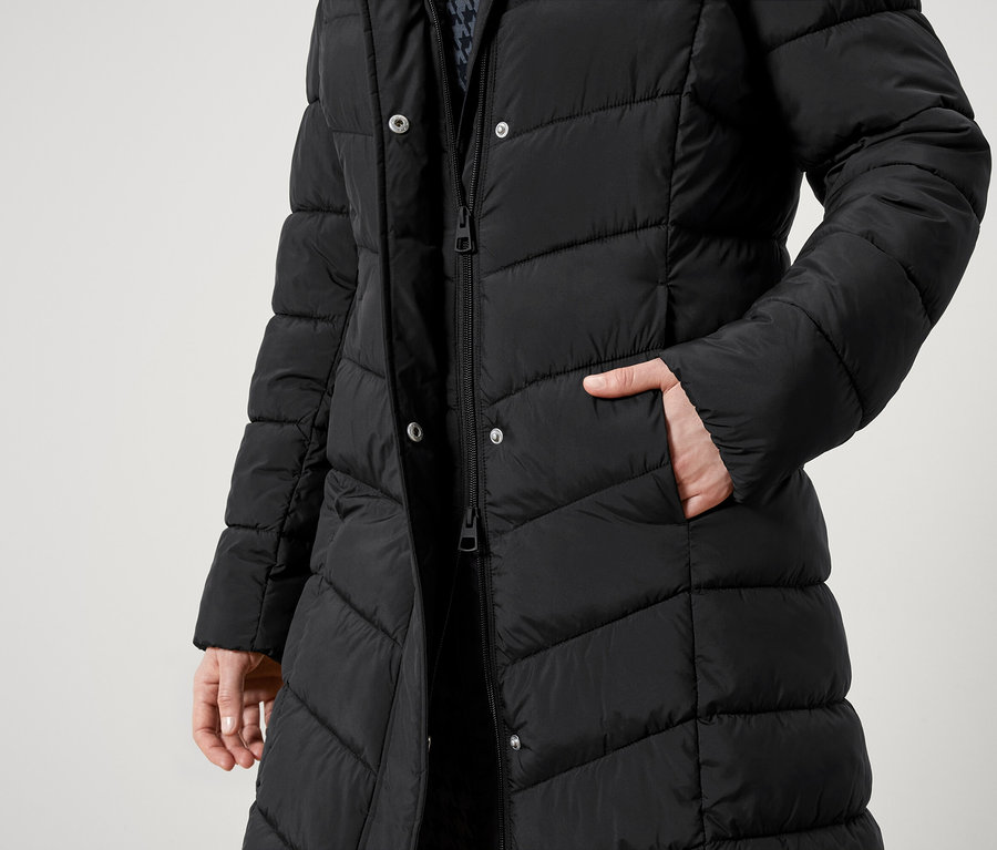 Teilansicht einer Person in einem schwarzen Steppmantel mit Reißverschluss und Knöpfen. Eine Hand steckt in der Tasche.