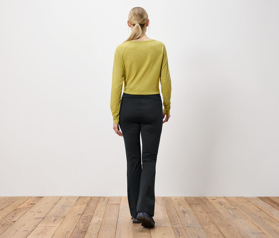 Frau mit blonden, zum Pferdeschwanz gebundenen Haaren steht mit dem Rücken zur Kamera und trägt ein olivgrünes Langarmshirt und eine schwarze Hose.