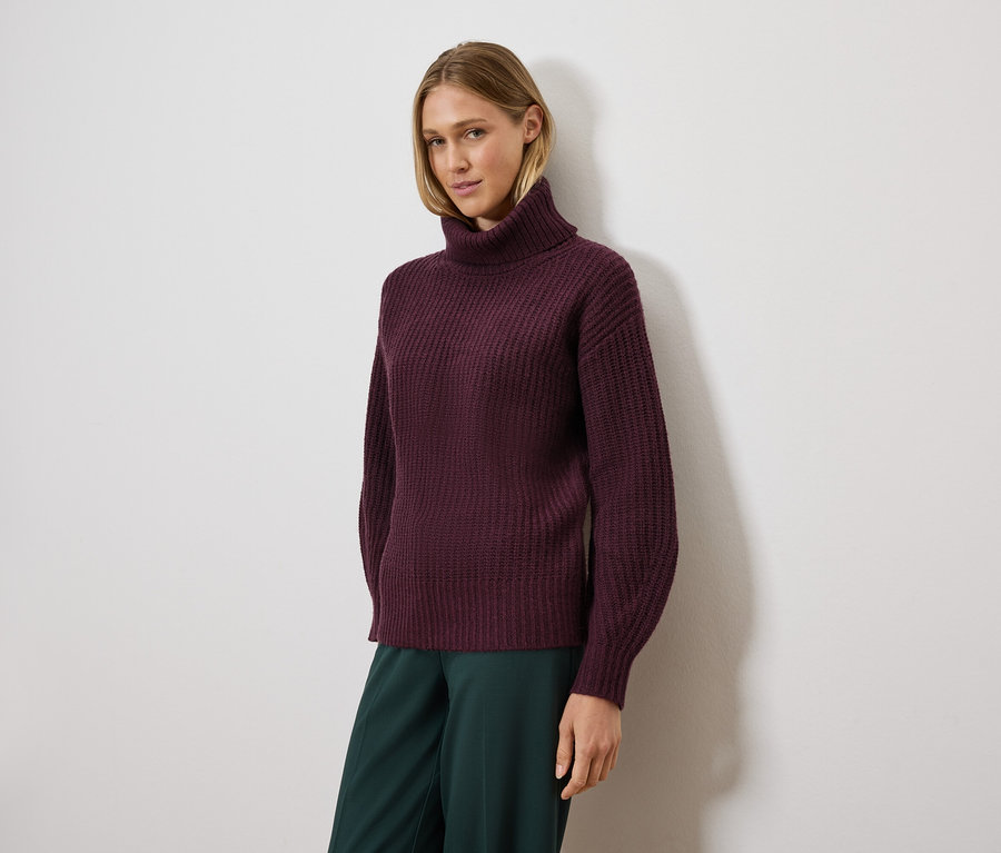 Frau posiert in einem violetten Rollkragenpullover und einer grünen Hose.