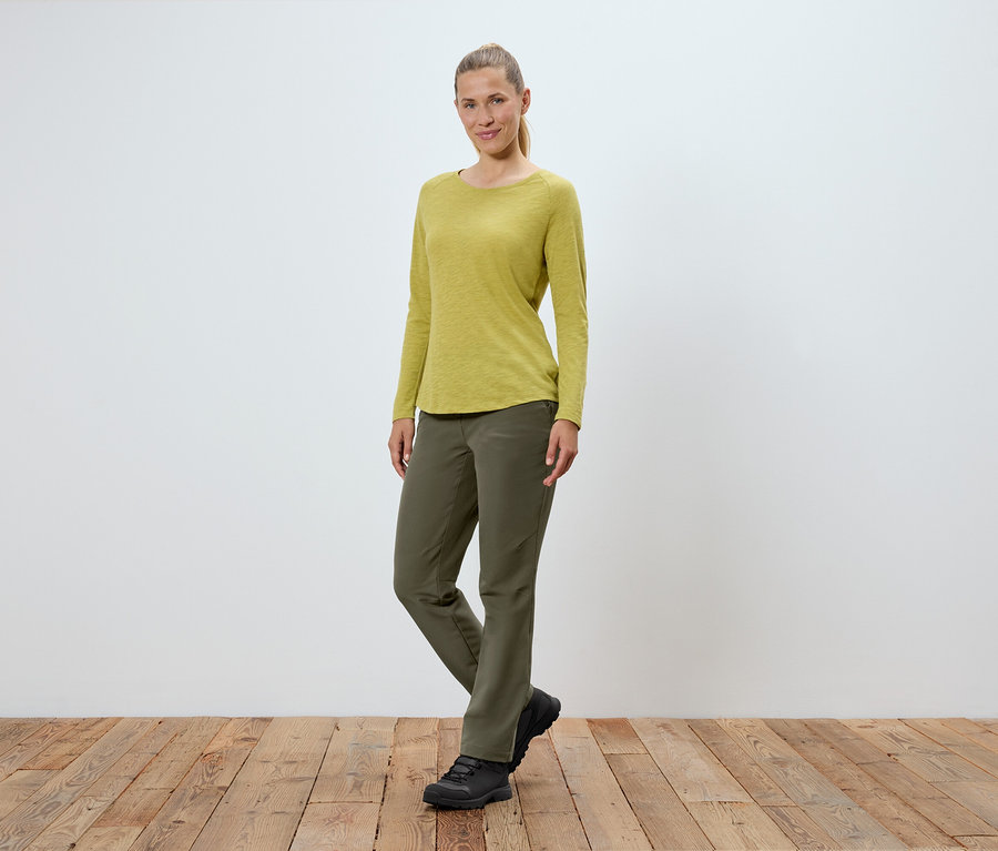 Eine Frau in olivgrüner Hose, gelbgrünem Langarmshirt und schwarzen Schuhen steht auf einem Holzboden vor einer weißen Wand.