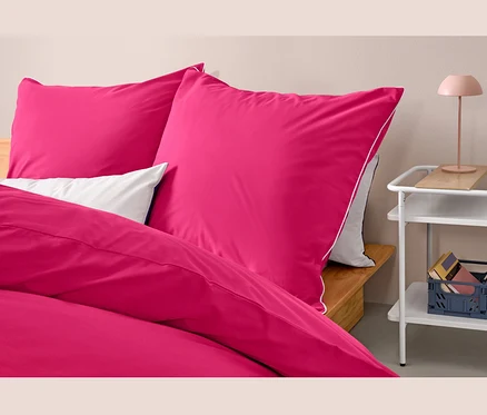 Nahaufnahme von drei Sets rosa Renforcé-Bettwäsche in Normalgröße auf einem Bett neben einem Tisch.