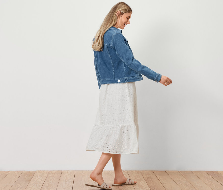 Frau geht, trägt eine Jeansjacke, ein Blusenkleid mit Lochstickerei und einen Shopper in Bastoptik.