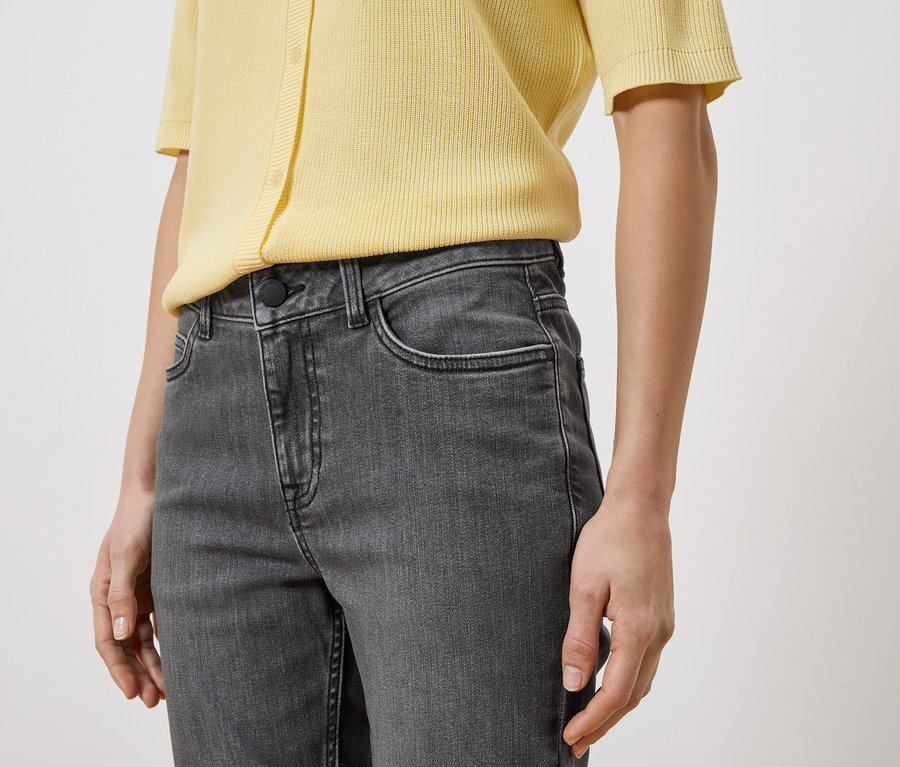 Nahaufnahme einer Person in grauen Jeans und einem gelben Oberteil vor einem weißen Hintergrund.