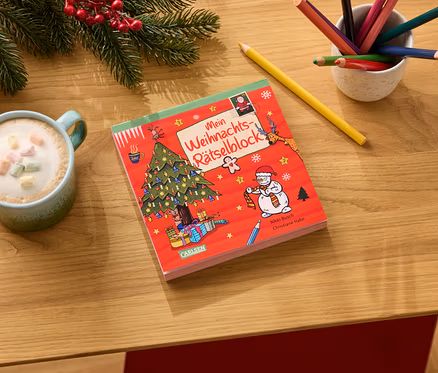 Auf einem Holztisch liegen ein roter Weihnachtsrätselblock, eine Tasse Kakao mit Marshmallows, Buntstifte und Tannenzweige.