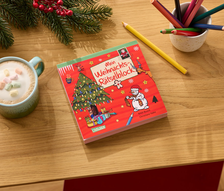 Auf einem Holztisch liegen ein roter Weihnachtsrätselblock, eine Tasse Kakao mit Marshmallows, Buntstifte und Tannenzweige.