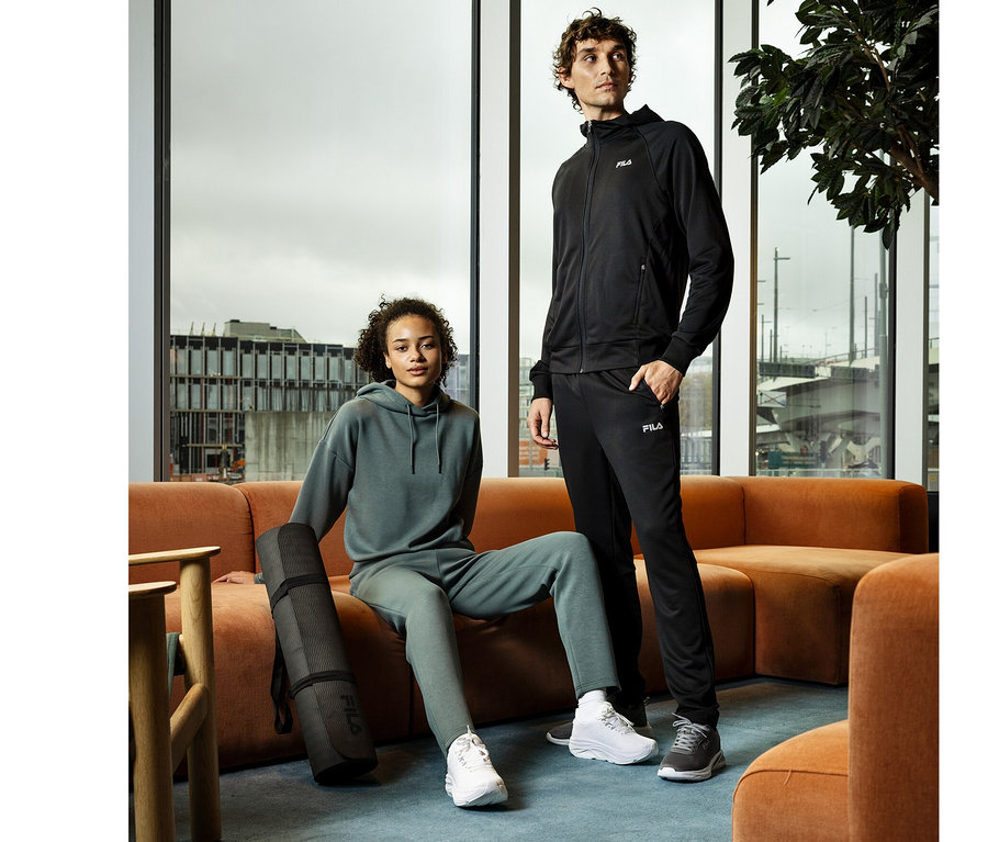 Eine Frau sitzt auf einem orangefarbenen Sofa neben einem stehenden Mann. Sie trägt einen grünen Hoodie und eine Jogginghose, daneben liegt eine schwarze Yogamatte. Er trägt einen schwarzen Hoodie und eine schwarze FILA Herren-Funktions-Sweathose.