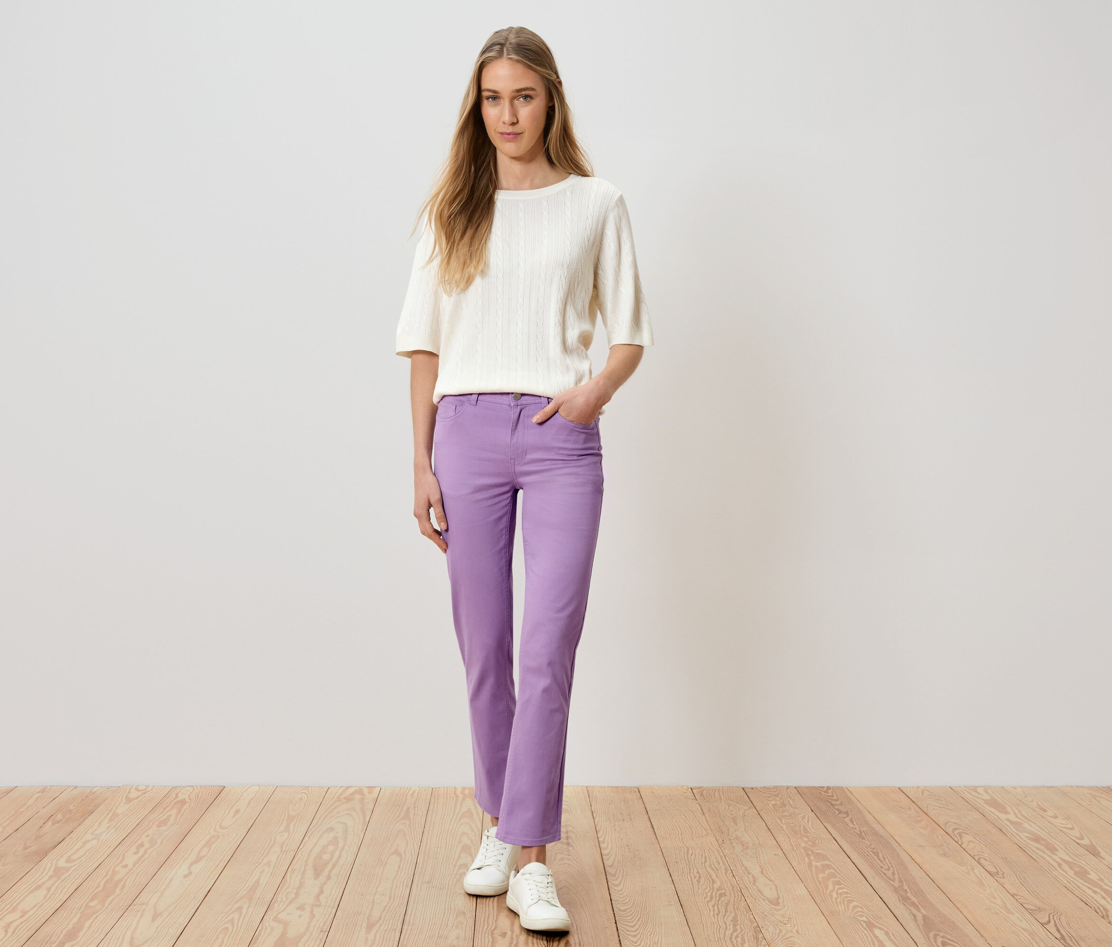 Frau steht mit Hand in Tasche und trägt lila Hose, weißen Pullover und weiße Schuhe.