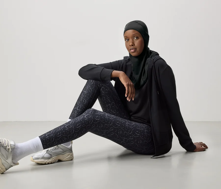 Frau mit schwarzem Hijab, Trainingsanzug und silbernen Turnschuhen sitzt auf dem Boden.