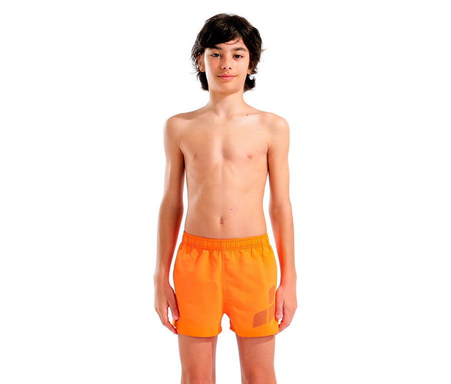 Ein barfüßiger Teenager steht vor einem weißen Hintergrund und trägt orangefarbene Badeshorts.