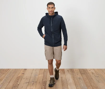 Ein Mann geht in Khaki-Shorts, einem dunkelblauen Kapuzenpullover und braunen Stiefeln auf einem Holzboden.