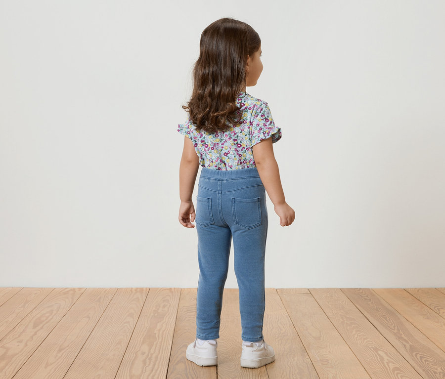 Rückansicht eines kleinen Mädchens mit lockigem Haar, das ein blaues, kurzärmeliges Blumenoberteil, blaue Leggings und weiße Turnschuhe trägt.