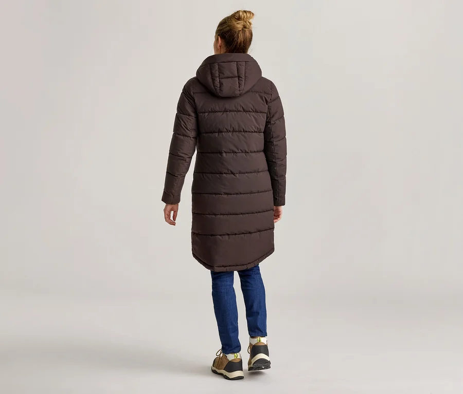Frau von hinten in braunem, langem Steppmantel mit Kapuze, blauen Jeans und braunen Schuhen vor hellem Hintergrund.