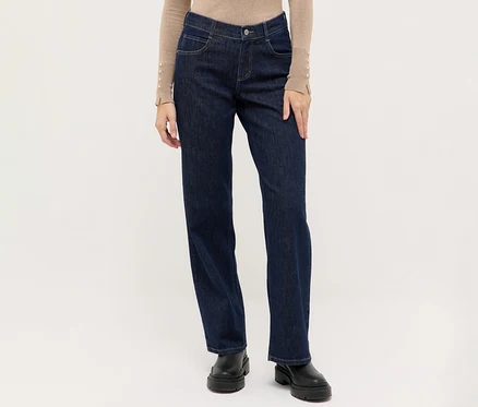 Person trägt dunkelblaue Jeans mit geradem Bein, ein beiges Langarmshirt mit Knopfdetails an den Ärmelbündchen und schwarze klobige Stiefel.