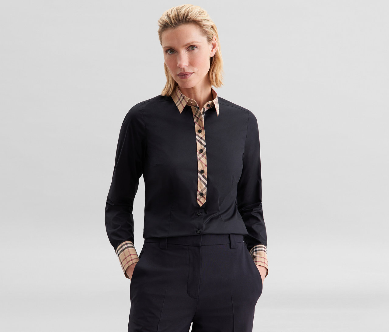 Frau trägt eine schwarze Seidensticker Popeline-Bluse mit Karodetail an Kragen, Knopfleiste und Manschetten, Hände in den Hosentaschen.