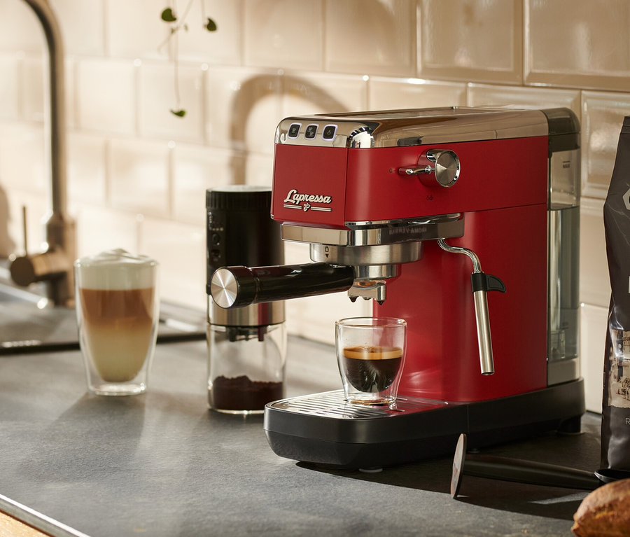 Eine rote »Lapressa« Siebträgermaschine steht auf einer Küchentheke neben einem Glas Kaffee.