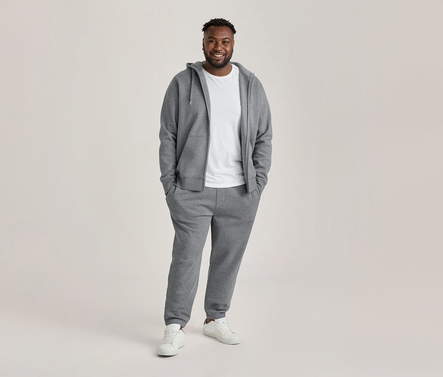 Lächelnder Mann trägt eine graue Kapuzenjacke mit Reißverschluss, ein weißes T-Shirt mit Rundhalsausschnitt, eine graue Jogginghose und weiße Sneaker, die Hände in den Taschen.
