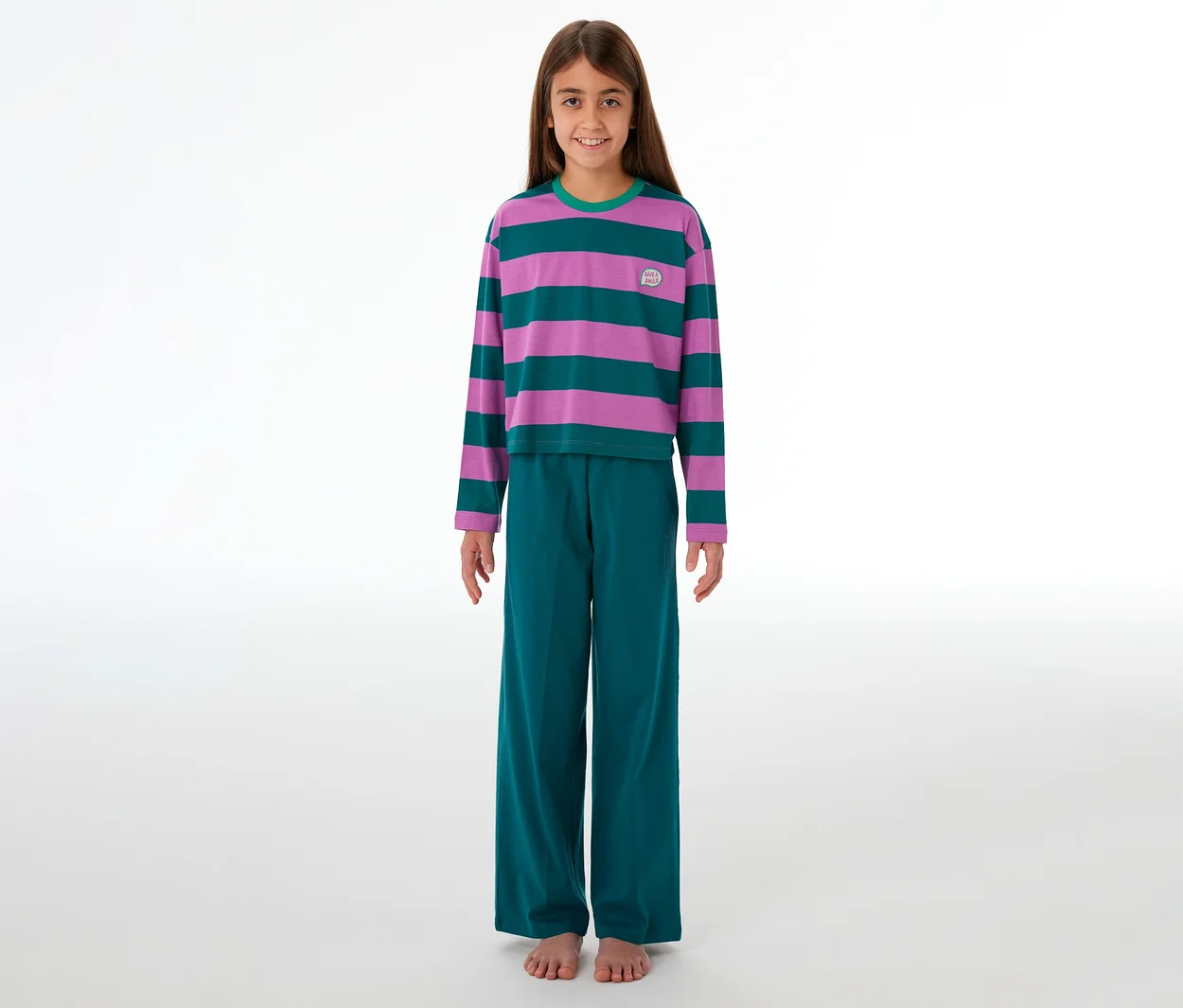 Ein junges Mädchen steht lächelnd barfuß in einem gestreiften SCHIESSER Kinder-Pyjama in Lila und Türkis mit passender Hose.