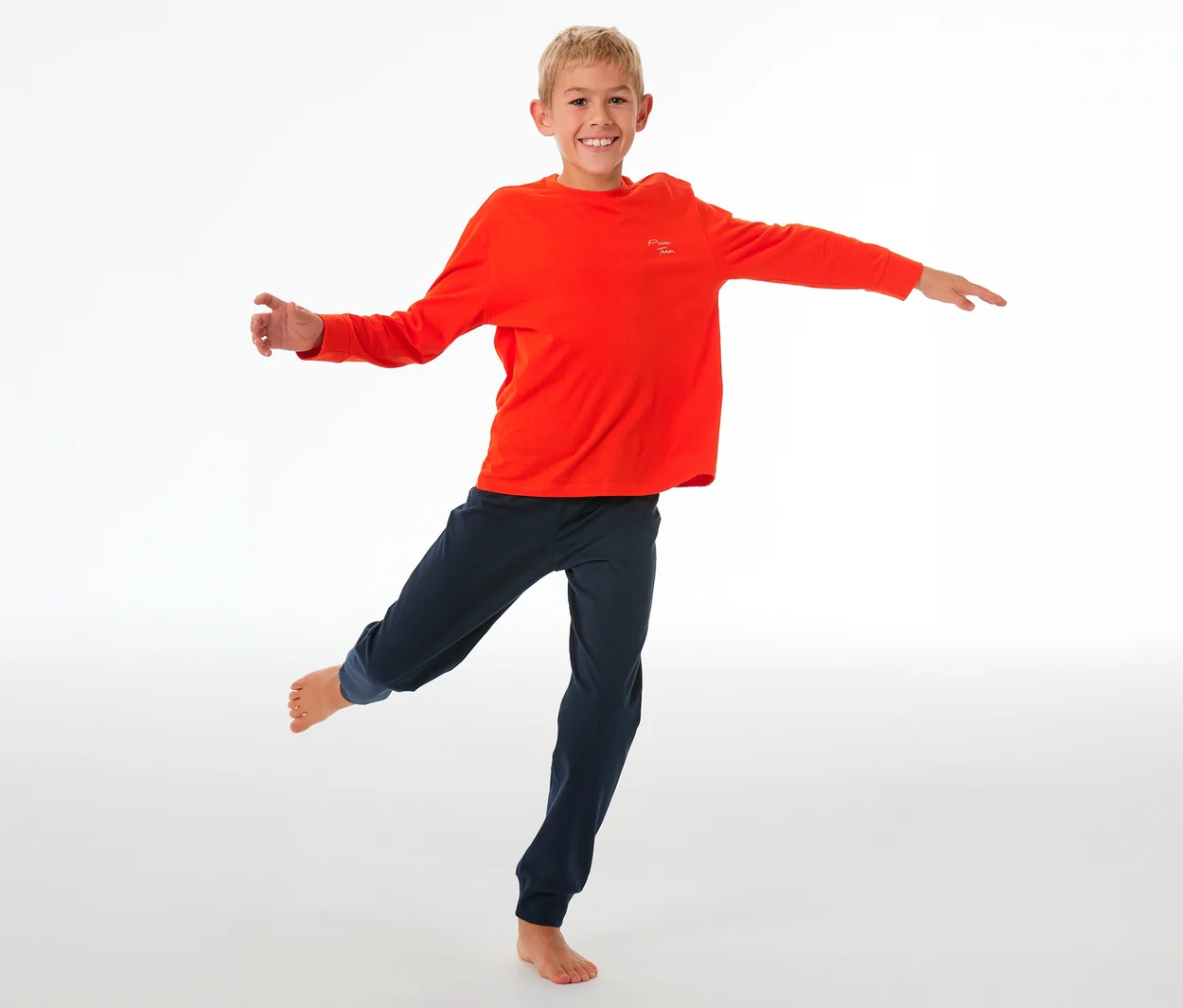 Lächelnder Junge balanciert auf einem Bein, trägt einen SCHIESSER Kinder-Pyjama mit Bündchen: orangefarbenes Langarmshirt und dunkelblaue Hose.