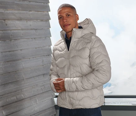 Mann posiert auf einem Balkon in beiger Steppjacke.