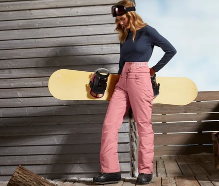 Frau steht an einer Holzwand und lehnt sich an ein Snowboard. Sie trägt eine Ski- und Snowboardhose.