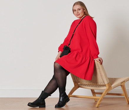 Frau sitzt auf einem Korbstuhl und trägt ein rotes Sweatkleid, Lederboots, 2 Paar Strumpfhosen und Mini-Taschen.