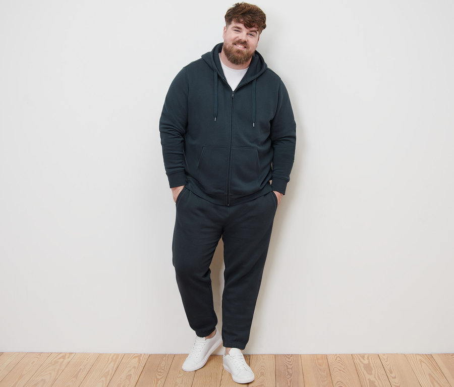 Ein Mann posiert in einer Kapuzenjacke, einer Sweathose und weißen Turnschuhen für die Kamera.