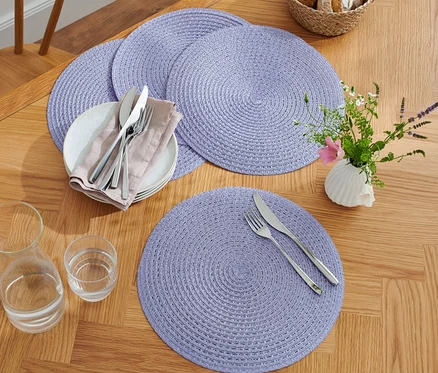 Vier lila Platzsets auf einem Holztisch mit Tellern, Besteck, Gläsern und einer Vase mit Blumen.