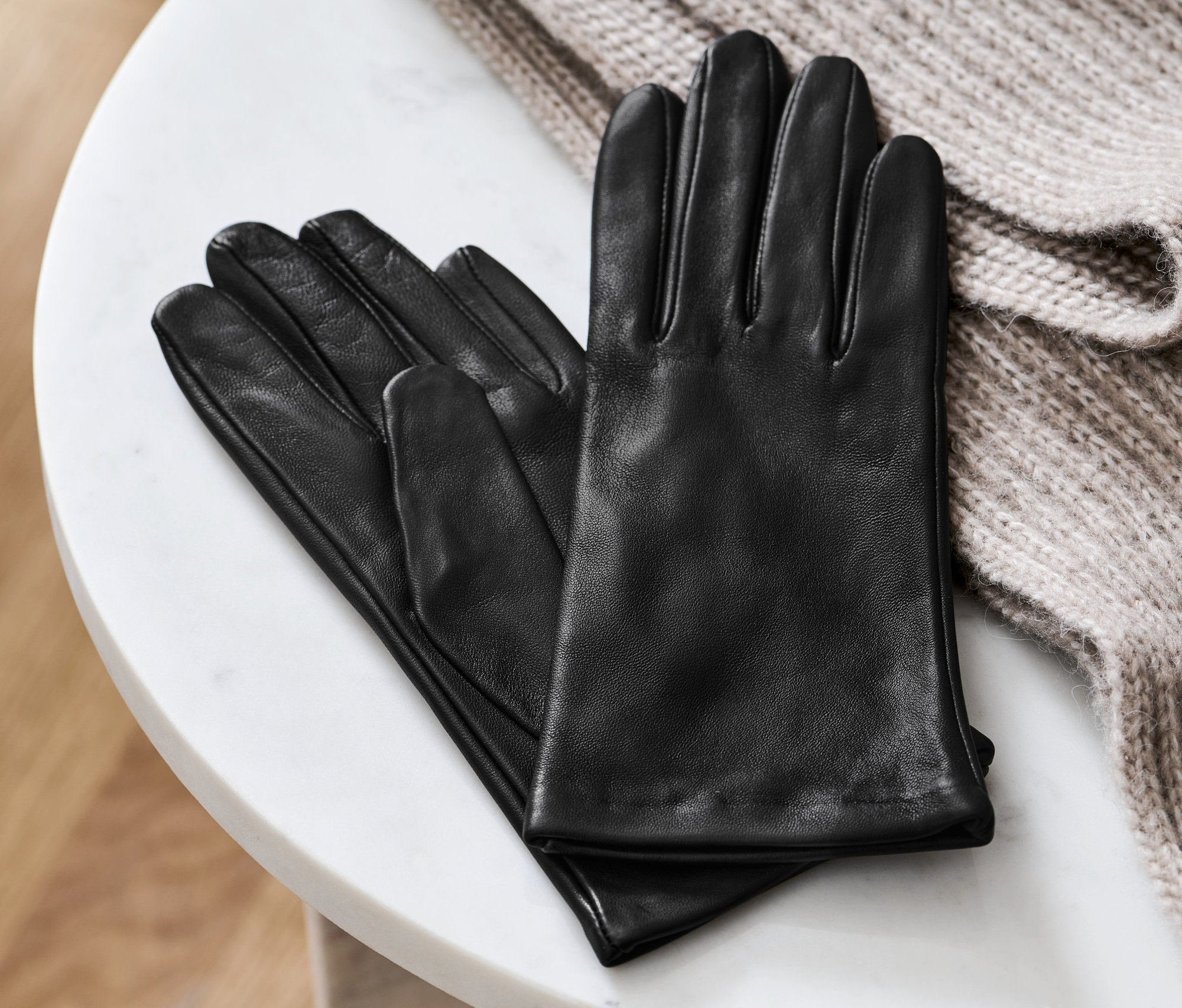 Schwarze Lederhandschuhe liegen auf einem weißen Marmortisch neben einem beigen Strickpullover.