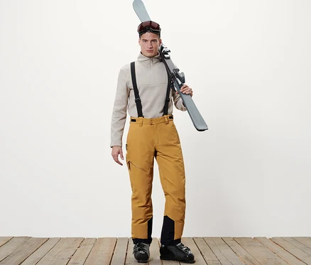 Mann steht mit Skiern über der Schulter, trägt einen beigen Pullover und eine Skihose in Camel.