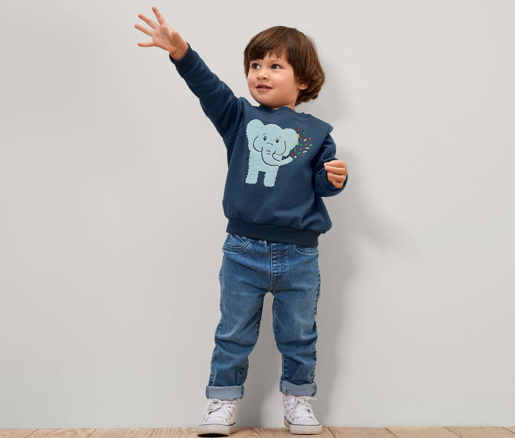 Ein Junge steht und winkt. Er trägt eine blaue Kinder-Pull-on-Pants – Fit »Luca« und einen blauen Pullover mit Elefant.