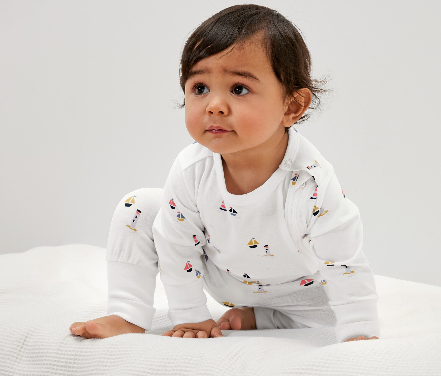 Baby in 3 maritimen, weißen Baby-Pyjamas sitzt auf weißem Stoff.