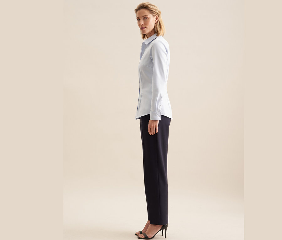 Frau posiert seitlich und trägt eine hellblaue Seidensticker Twillbluse, eine schwarze Hose und schwarze Schuhe mit Absatz.
