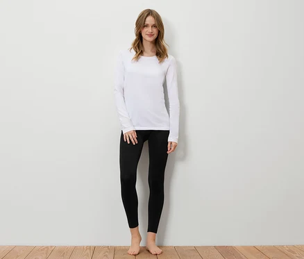 Frau steht vor einer weißen Wand, trägt ein weißes Langarmshirt und schwarze Leggings.