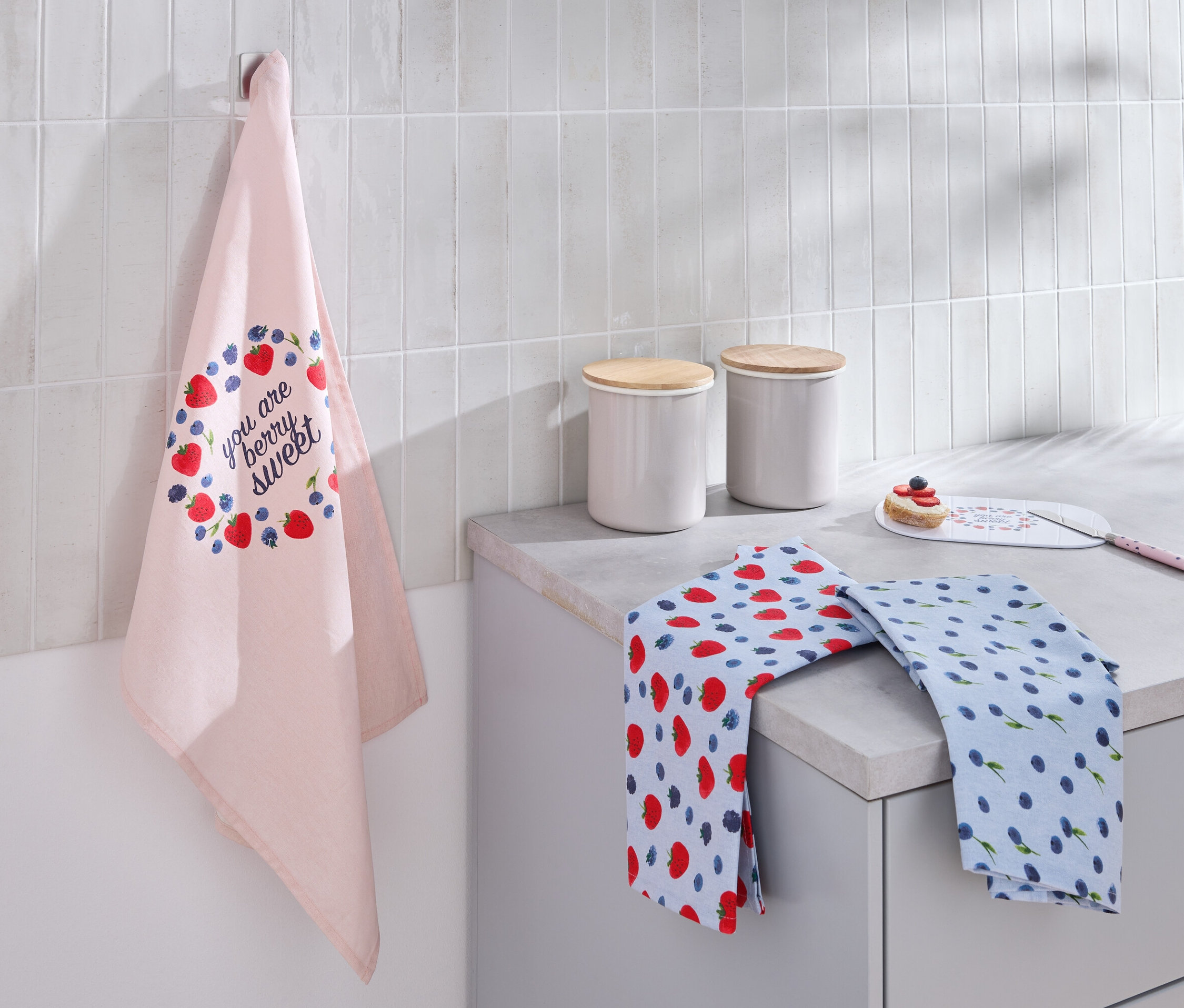 Ein hellrosa Geschirrtuch mit Erdbeer- und Blaubeeraufdruck hängt an der Wand. Auf der Arbeitsfläche liegen zwei gefaltete Geschirrtücher und zwei weiße Kanister mit Holzdeckeln.