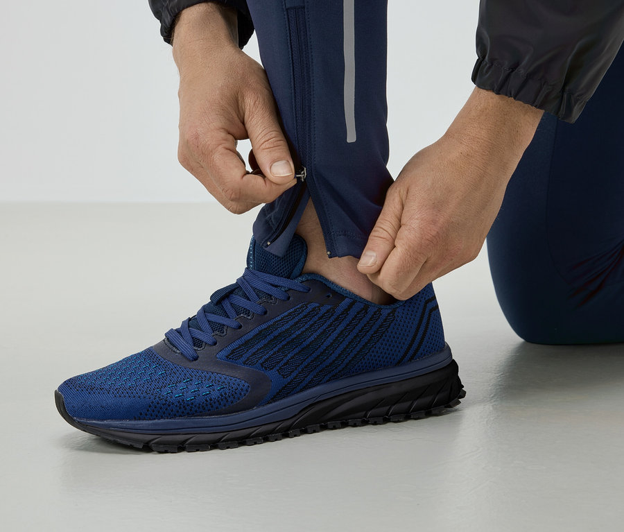 Mann schließt den Reißverschluss an einer blauen Laufhose, trägt blaue Schuhe.