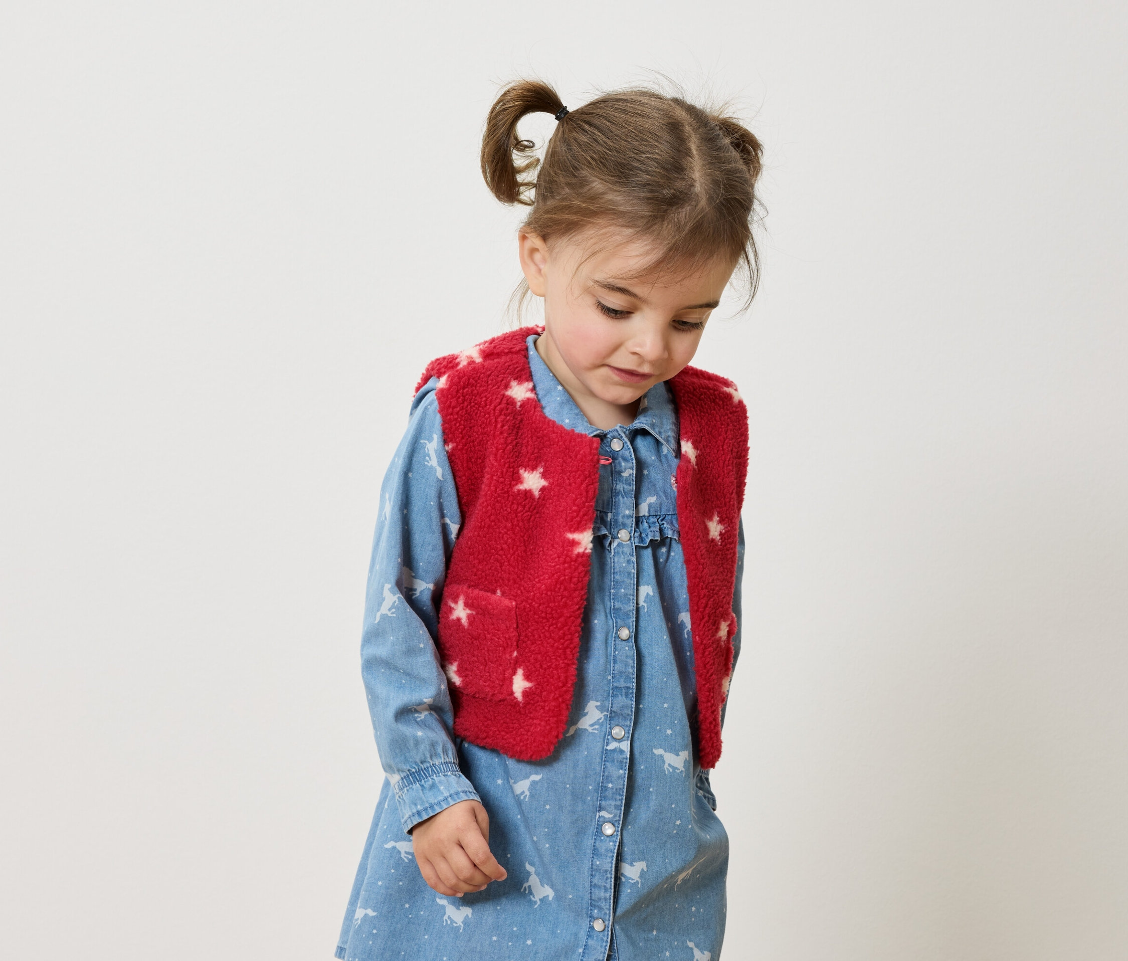 Mädchen mit braunen Zöpfen trägt ein blaues Kleid mit Pferdeaufdruck und eine rote Weste mit Sternen.