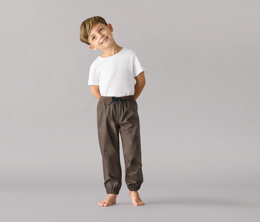 Ein Junge in einem weißen T-Shirt und einer Kinder-Pull-on-Hose – Fit »Anton« steht barfuß vor einem grauen Hintergrund.