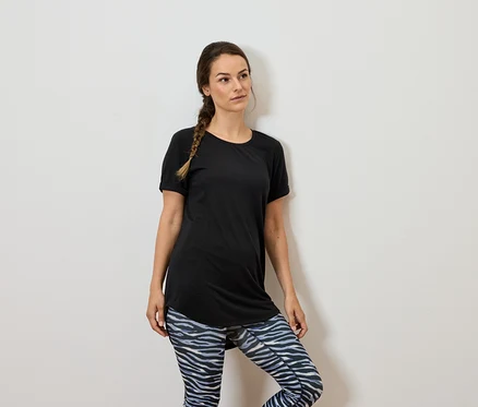Frau mit braunen Haaren in schwarzem T-Shirt und gemusterten Leggings steht an einer weißen Wand.