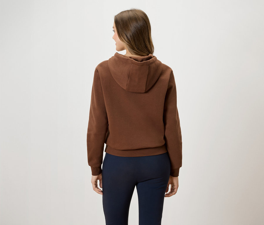 Rückansicht einer Frau mit braunen Haaren, die einen braunen Kapuzenpullover und eine dunkelblaue Hose trägt.