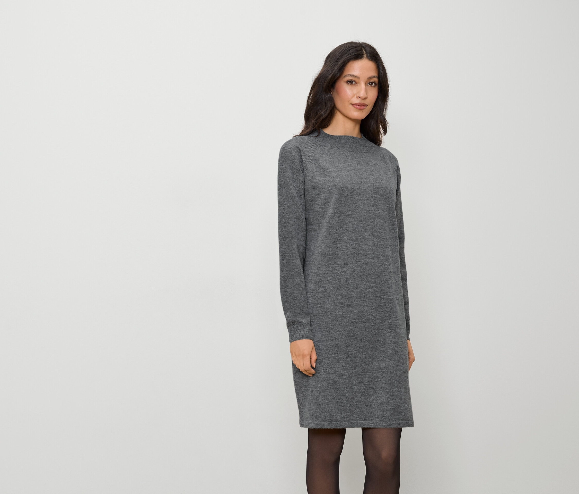 Eine Frau in einem langen, grauen Kleid und schwarzer Strumpfhose steht vor einem weißen Hintergrund.