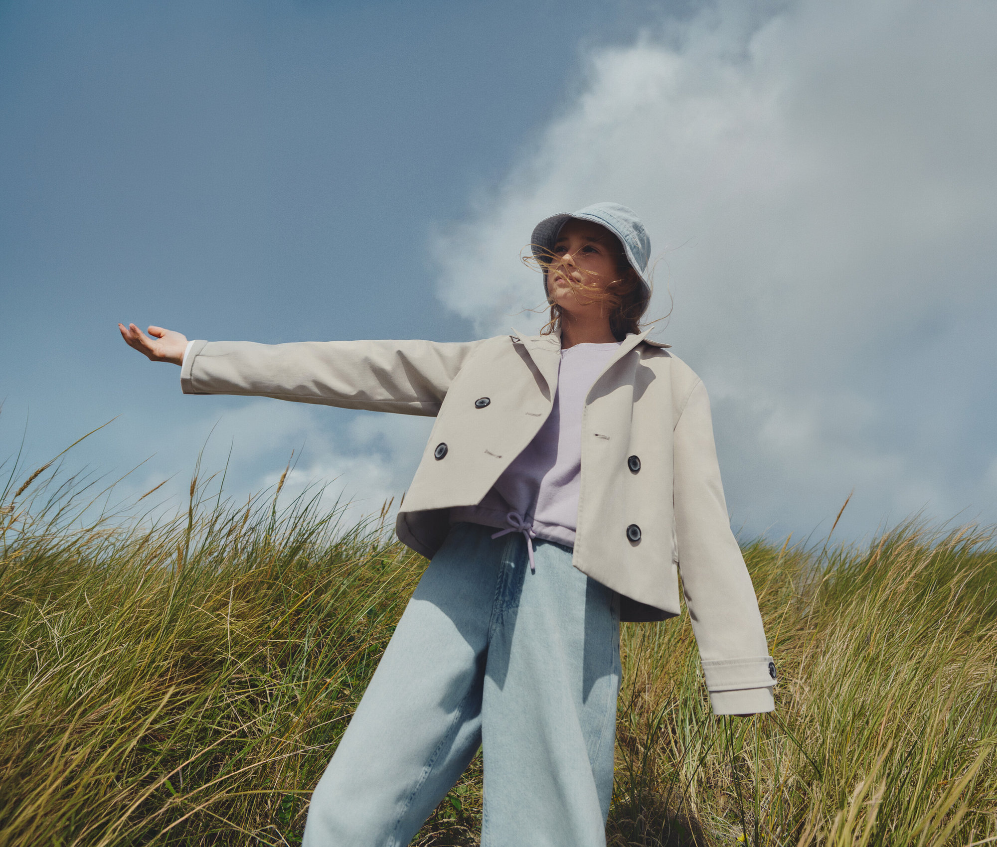 Eine junge Frau steht in einem Feld und trägt einen hellbraunen Trenchcoat, ein lila Sweatshirt, Jeans und einen blauen Fischerhut.