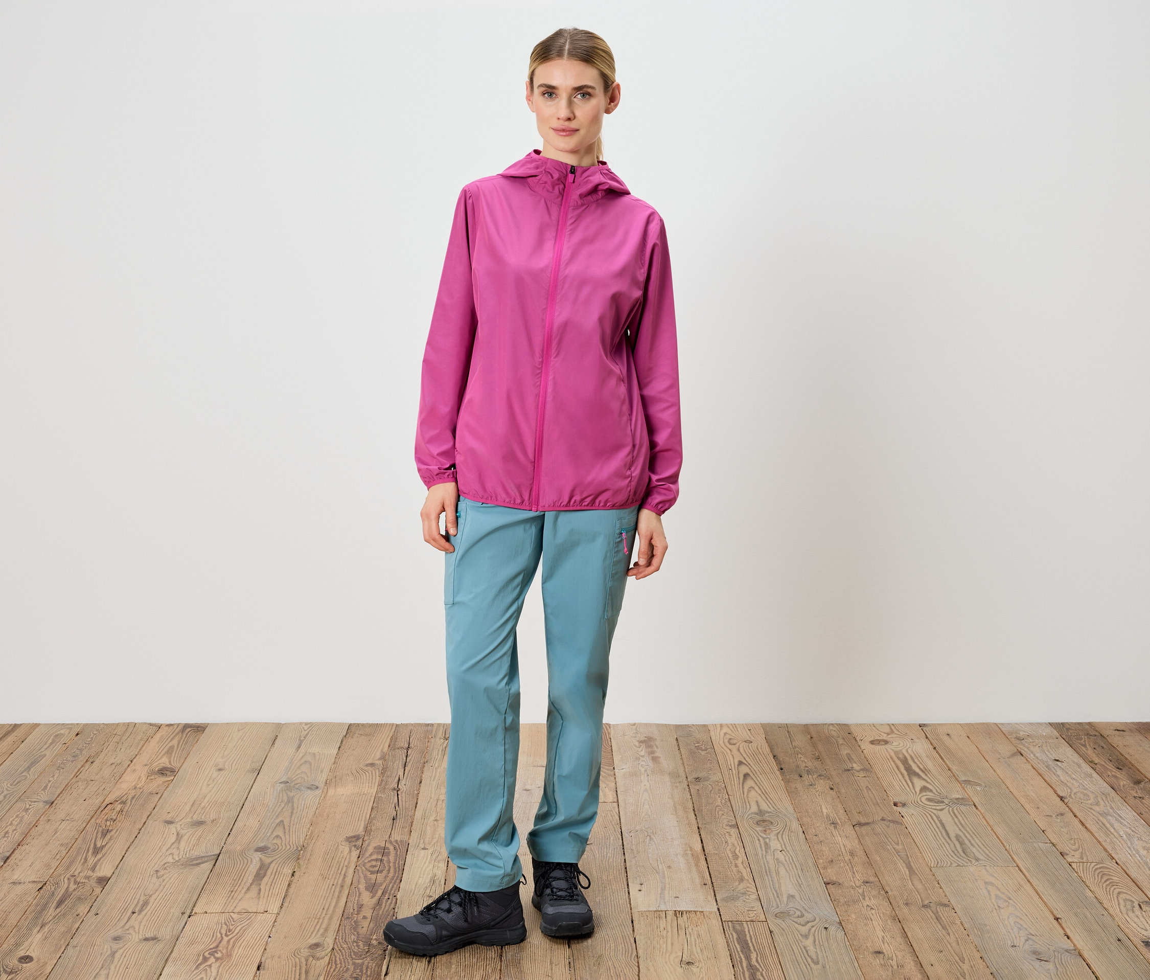 Frau steht vor einer weißen Wand und trägt eine pinkfarbene Jacke, blaue Hose und schwarze Schuhe.