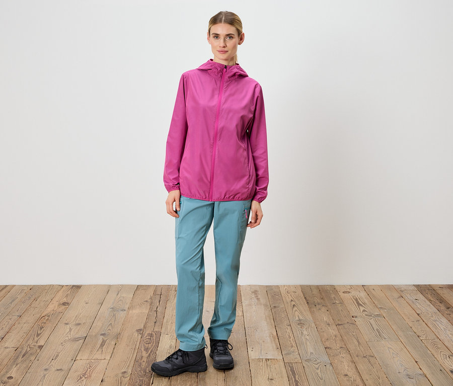 Frau steht vor einer weißen Wand und trägt eine pinkfarbene Jacke, blaue Hose und schwarze Schuhe.