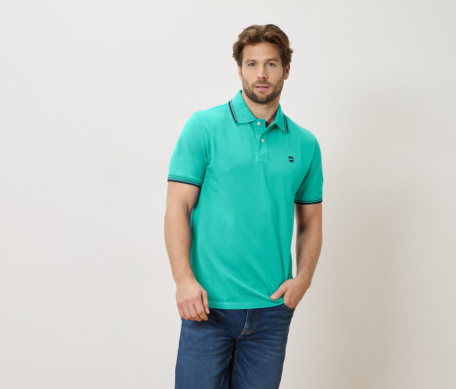 Mann mit einem smaragdgrünen Poloshirt und Jeans, eine Hand in der Hosentasche.