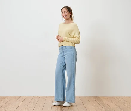 Frau mit Pferdeschwanz, gelbem Pullover, hellblauen Jeans und weißen Schuhen steht vor einer weißen Wand auf Holzboden und lächelt.