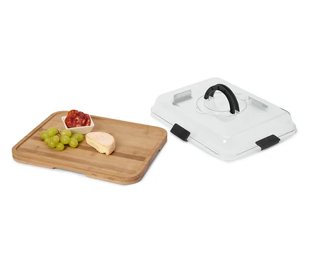 Käse, Wurst und Weintrauben auf einem Holzbrett neben einer transparenten Servierhaube mit schwarzem Griff.