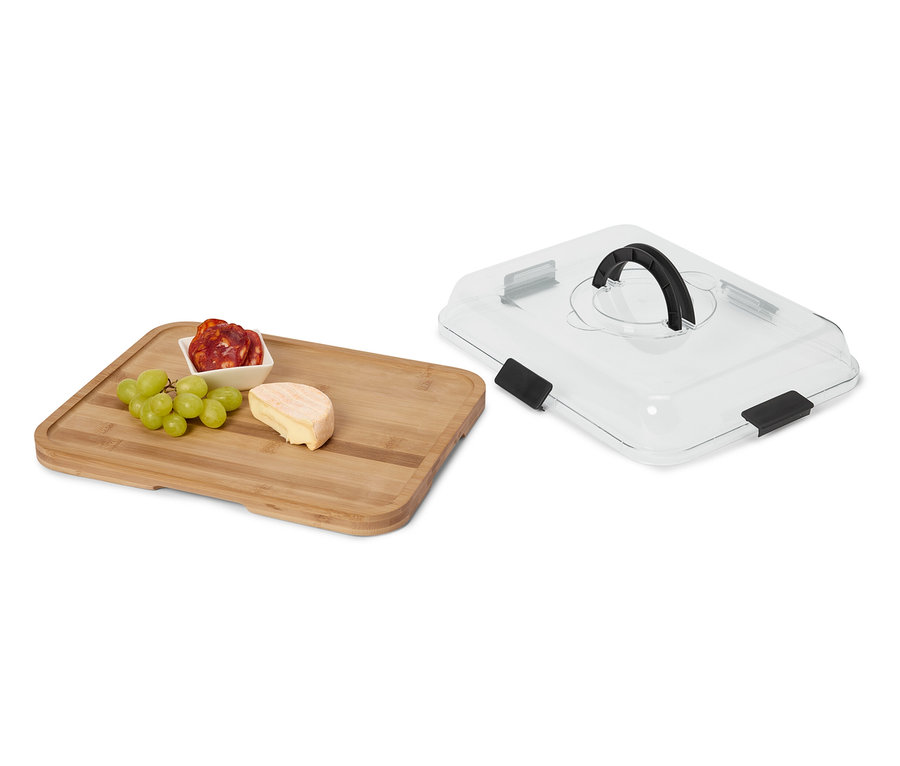 Käse, Wurst und Weintrauben auf einem Holzbrett neben einer transparenten Servierhaube mit schwarzem Griff.