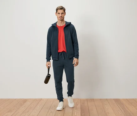 Mann steht in marineblauem Trainingsanzug und rotem T-Shirt, hält eine Trinkflasche in der Hand.
